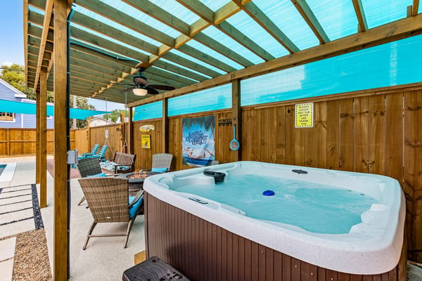 Covered patio with hot tub, wicker chairs, ceiling fan, and turquoise shade panels overlooking backyard