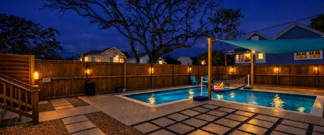 Backyard pool area at dusk with two illuminated pools, wooden fence, patio pavers, and neighboring houses under blue twilight sky