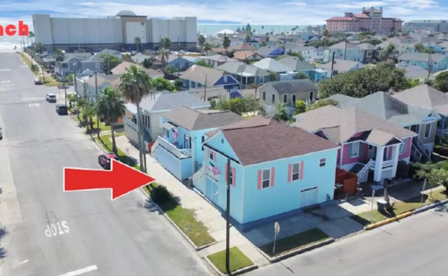 Aerial view of turquoise and pink cottage with red arrow pointing to it, surrounded by colorful houses and ocean in background